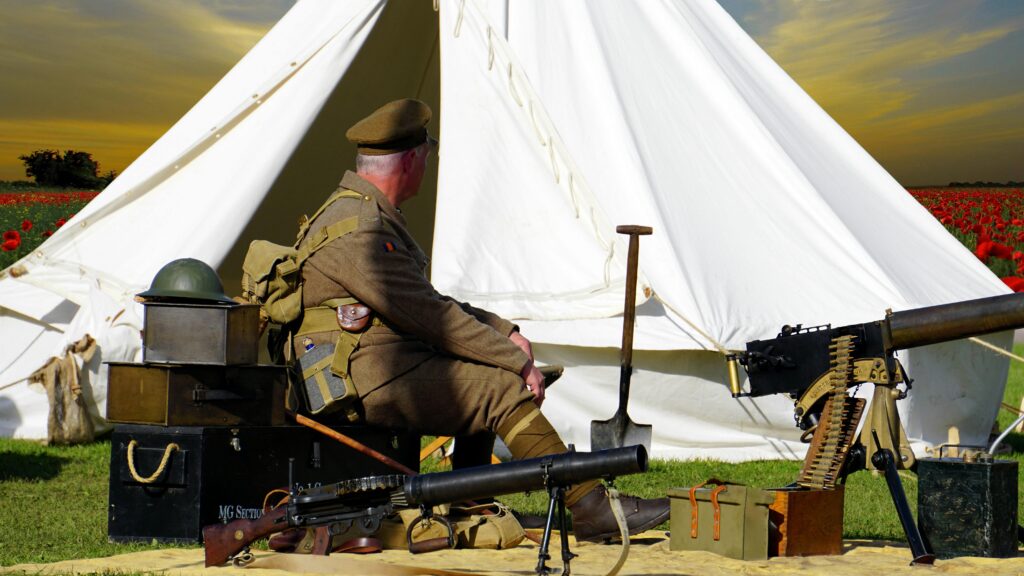 A historical reenactor dressed as a WWI soldier sits by a tent with military gear in a field.